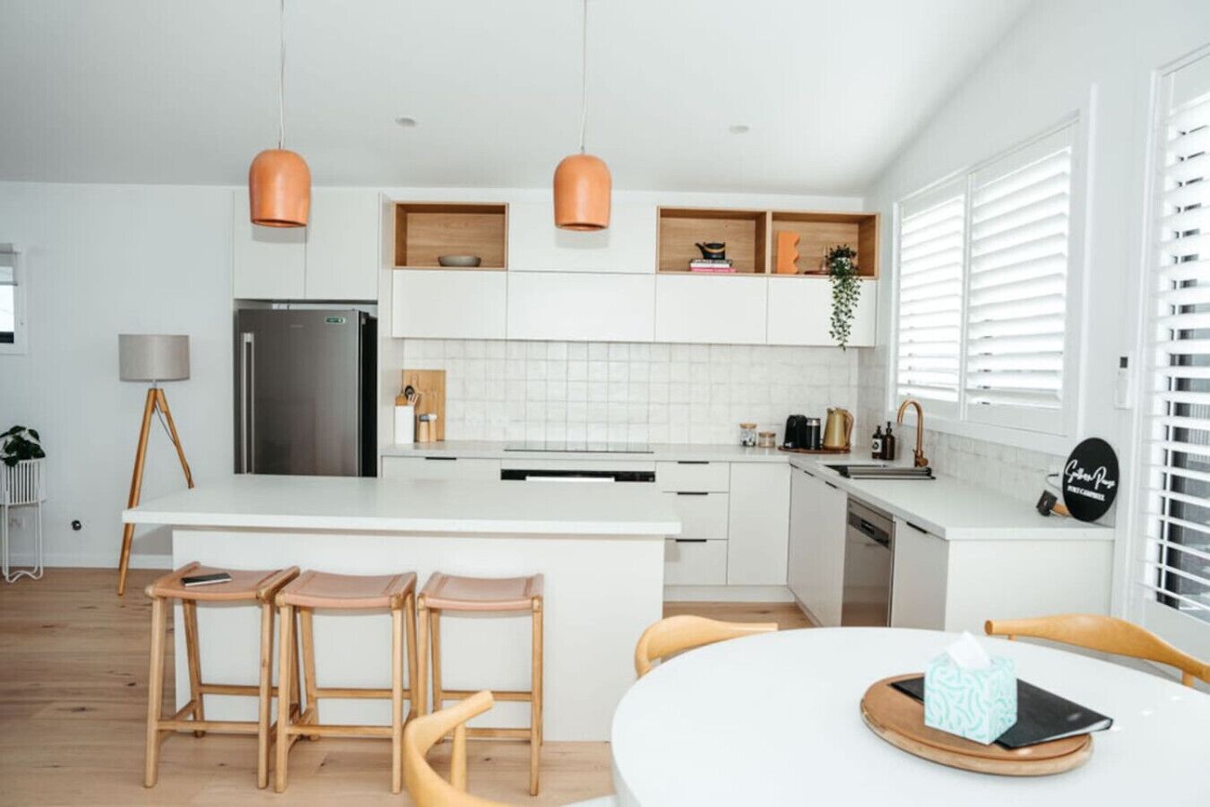 A modern kitchen with white cabinets, a central island with bar stools, pendant lighting, and a dining area with a table and chairs.