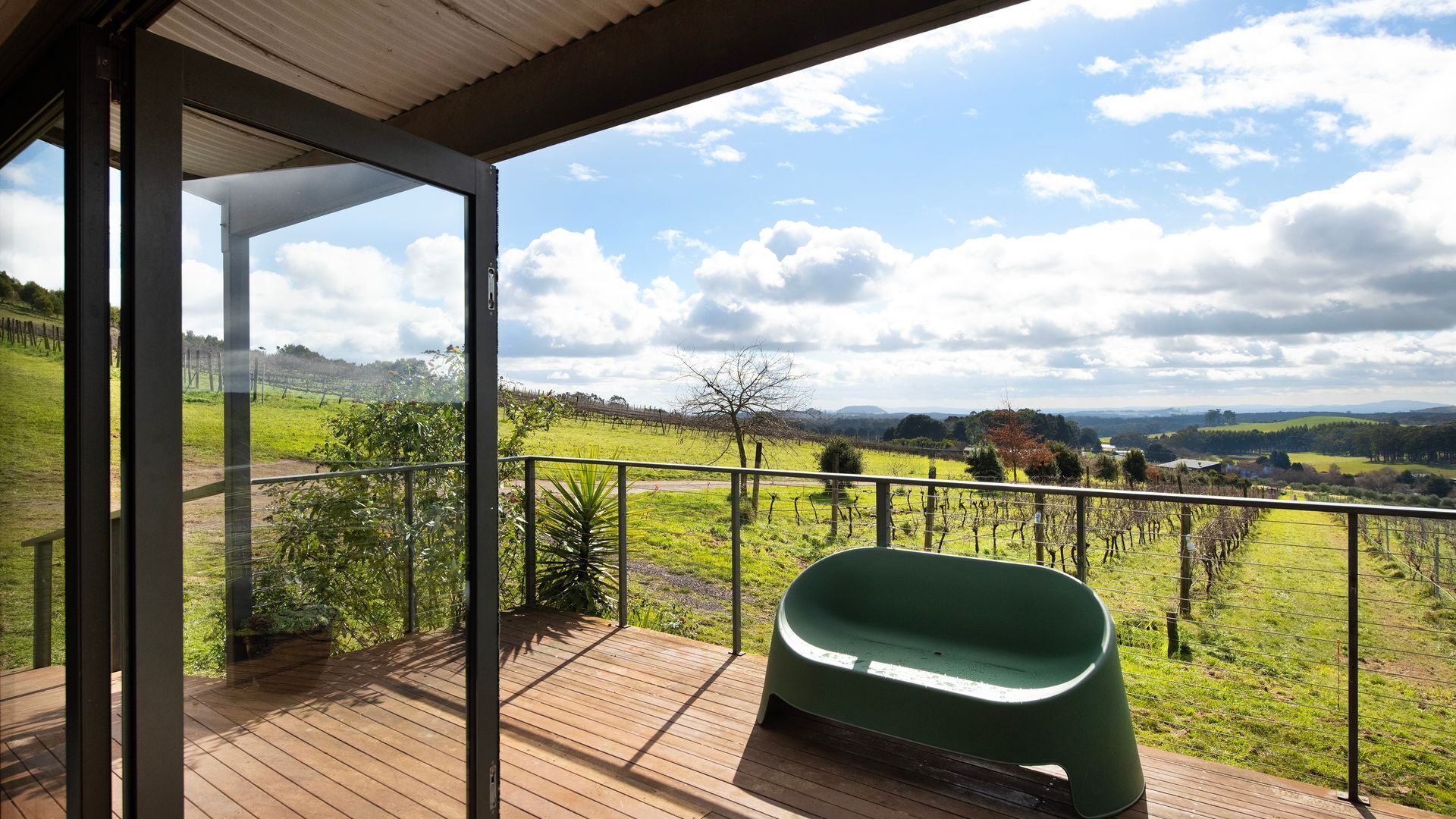 A balcony with a green plastic chair, wooden deck, and a view of a vineyard and hills.