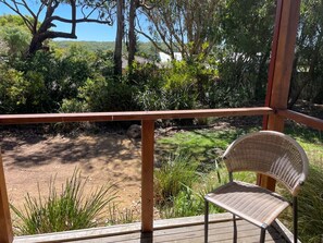 Una terraza de madera con una silla de mimbre, con vistas a un sendero de tierra y vegetación.
