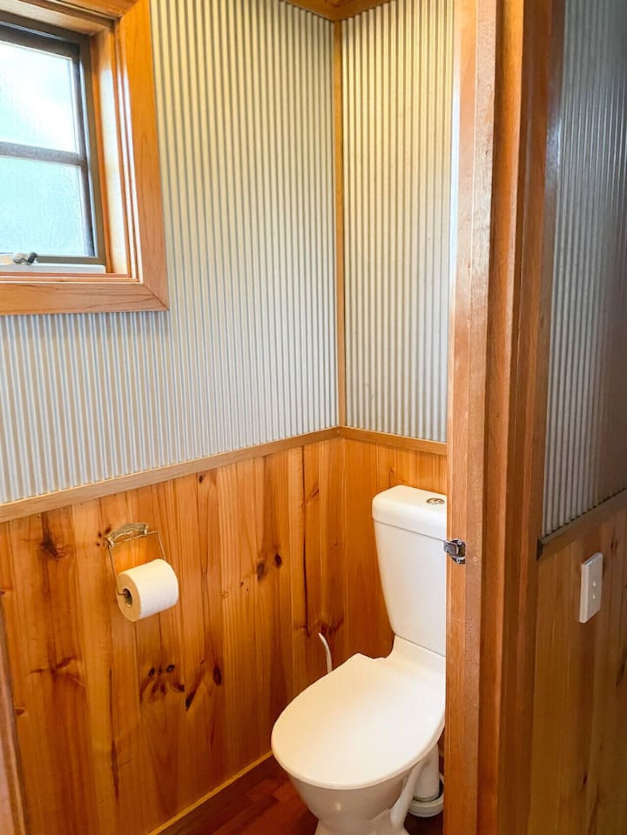 A small, compact bathroom with wooden paneling, a toilet, and a window.
