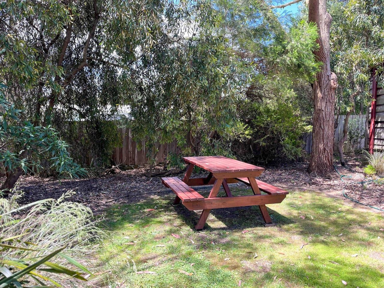 A wooden picnic table in a garden with dense foliage and a wooden fence in the background.