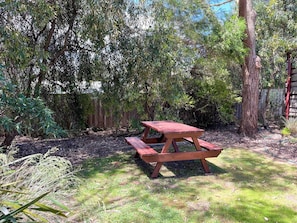 Una taula de picnic de fusta en un jardí amb vegetació abundant i una tanca de fusta al fons.