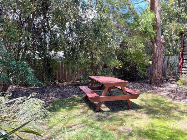 A wooden picnic table in a garden with dense foliage and a wooden fence in the background.