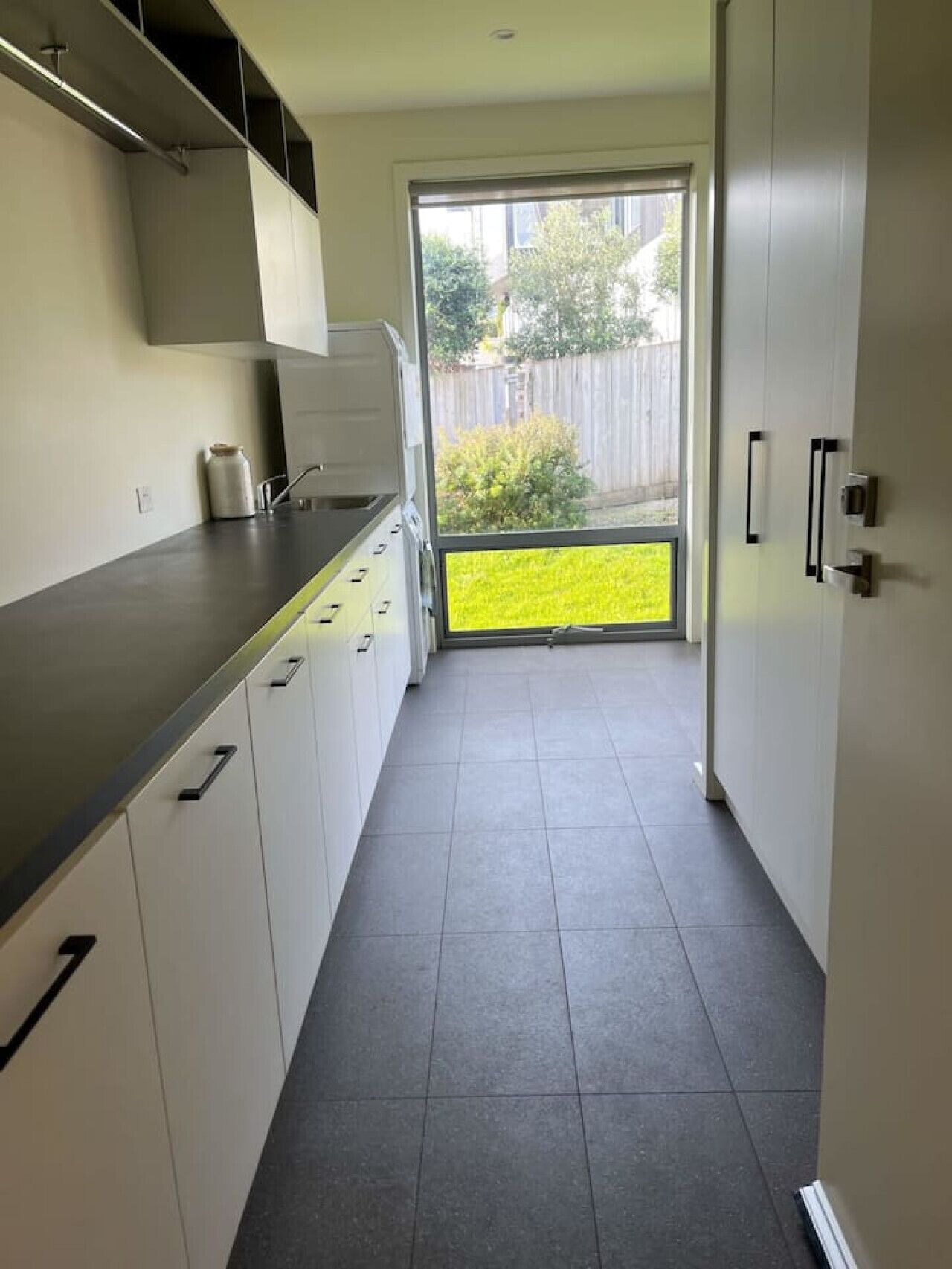 A narrow kitchen with white cabinets, a dark countertop, and a view of a backyard through a large window.