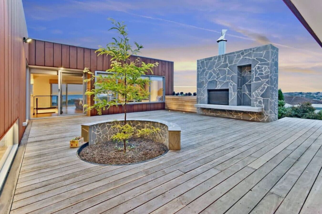 A modern outdoor deck with a stone fireplace, a small tree, and a view of a cityscape.