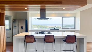 A modern kitchen with a large island, three bar stools, and a view of the sea.