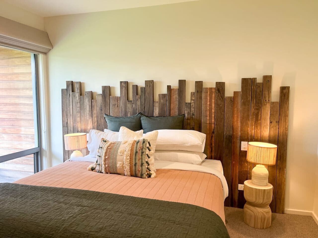 A bedroom with a wooden headboard, a bed with multiple pillows, two bedside lamps, and a window with blinds.