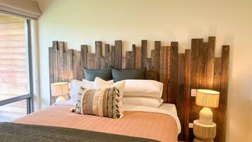 A bedroom with a wooden headboard, a bed with multiple pillows, two bedside lamps, and a window with blinds.
