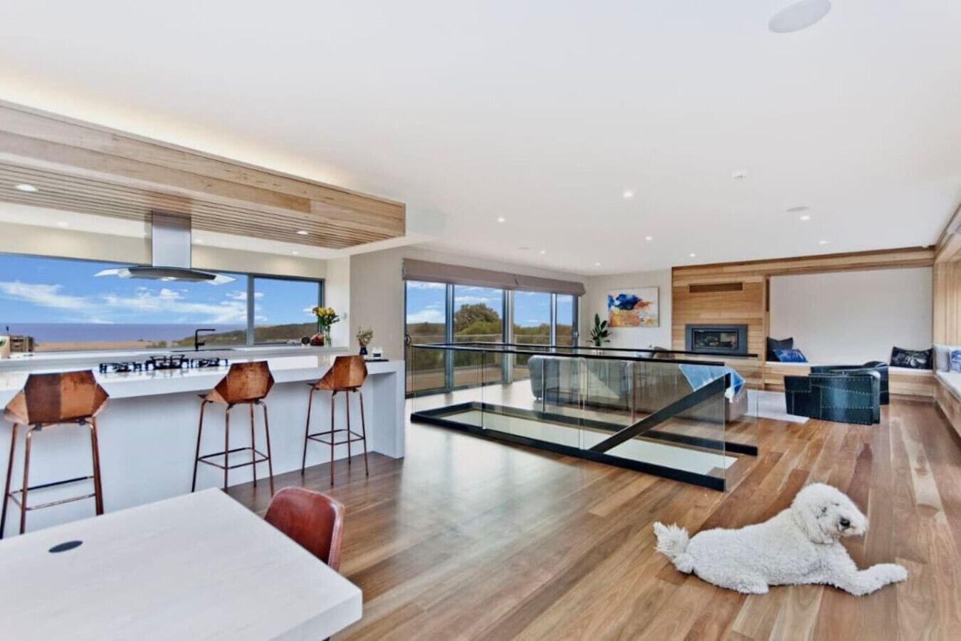 A modern interior with a kitchen, dining area, and a living space with a glass railing overlooking a scenic view.