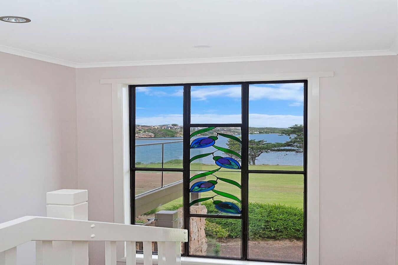 A room with a large window featuring a decorative glass design, a white railing, and a view of a lake and distant landscape.