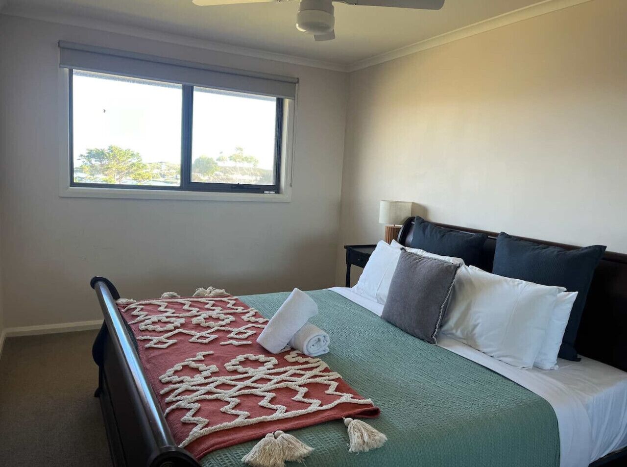 Una cama bien hecha con una colcha estampada, almohadas y una toalla enrollada. Se ve una ventana con persianas, una mesita de noche con una lámpara y un ventilador de techo.