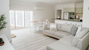 A modern living room with a sofa, dining table, and chairs. There is a kitchen area with cabinets and appliances.