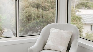 A corner room with a grey armchair and a white cushion, large windows with a view of trees and buildings.