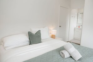 A hotel room with a bed, pillows, a bedside lamp, and a bathroom visible in the background. - Port Campbell Coastal Unit 3 (Port Campbell)