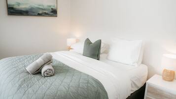 A neatly made bed with a quilted green blanket, two pillows, and rolled towels. There is a bedside lamp on the right and a painting on the wall above the bed.