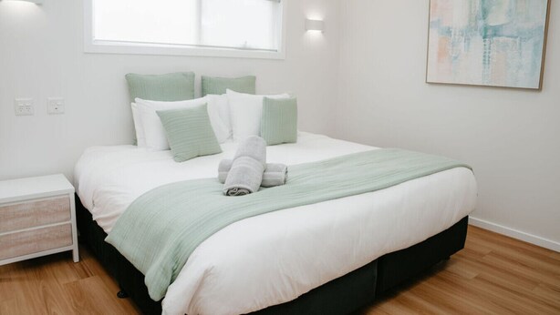 A neatly made bed with a green blanket and pillows, a bedside table, a wall-mounted light, and a painting on the wall.