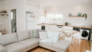 A modern living room with a beige sectional sofa, a dining table with chairs, a kitchen area with white cabinets, a washing machine, and a window providing natural light.