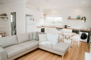 A modern living room with a beige sectional sofa, a dining table with chairs, a kitchen area with white cabinets, a washing machine, and a window providing natural light.