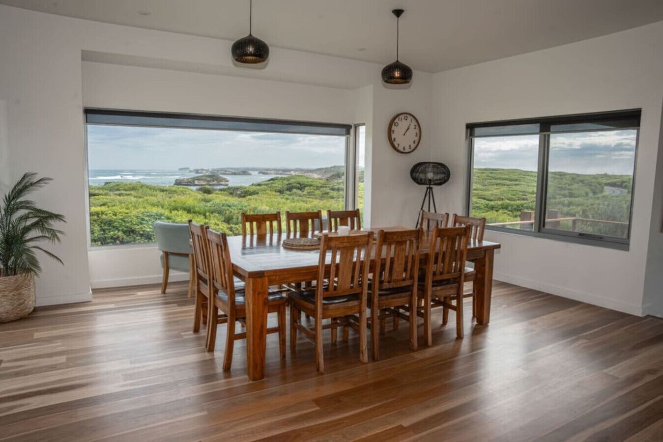 A dining area with a wooden table and chairs, large windows with a view of greenery and water, and a clock on the wall.