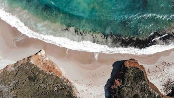 An aerial view of a beach with clear turquoise waters, white sandy shore, and rocky outcrops.