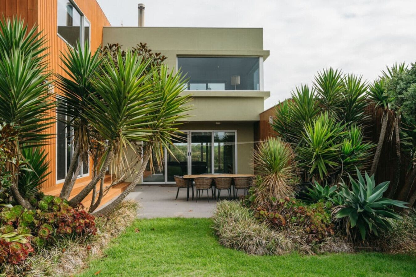 A modern two-story house with a patio area, surrounded by tropical plants.