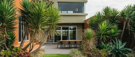 A modern two-story house with a patio area, surrounded by tropical plants.