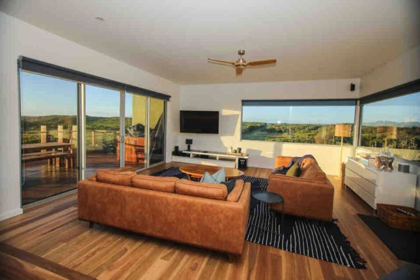 A modern living room with a large sofa, a flat-screen TV, and a dining area with a view of the outdoors.