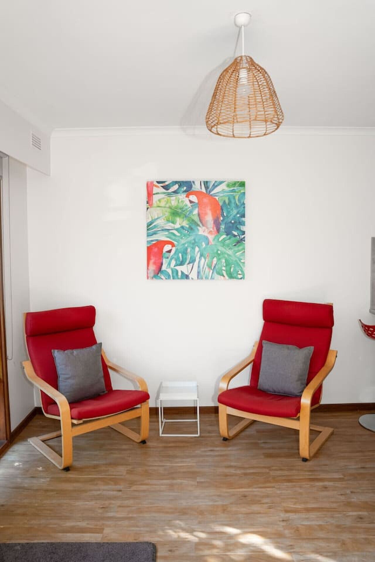 A room with two red chairs, a white table, and a painting of tropical birds on the wall.