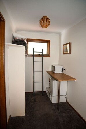 Une petite pièce avec un réfrigérateur blanc, un bureau en bois sur lequel se trouve un micro-ondes, une échelle et une fenêtre avec un cadre en bois.