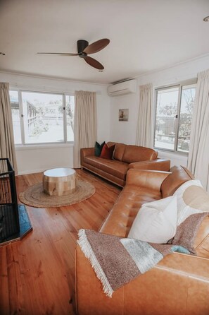 A living room with a brown leather sofa, a round wooden coffee table, and a ceiling fan. - Coastaways Port Campbell (Port Campbell)