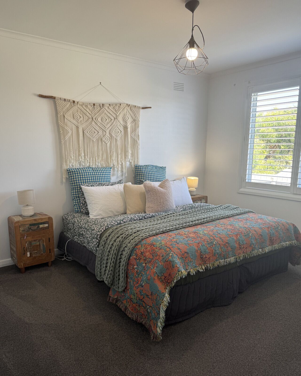 A bedroom with a bed, a macrame wall hanging, a hanging light fixture, and a window with blinds.