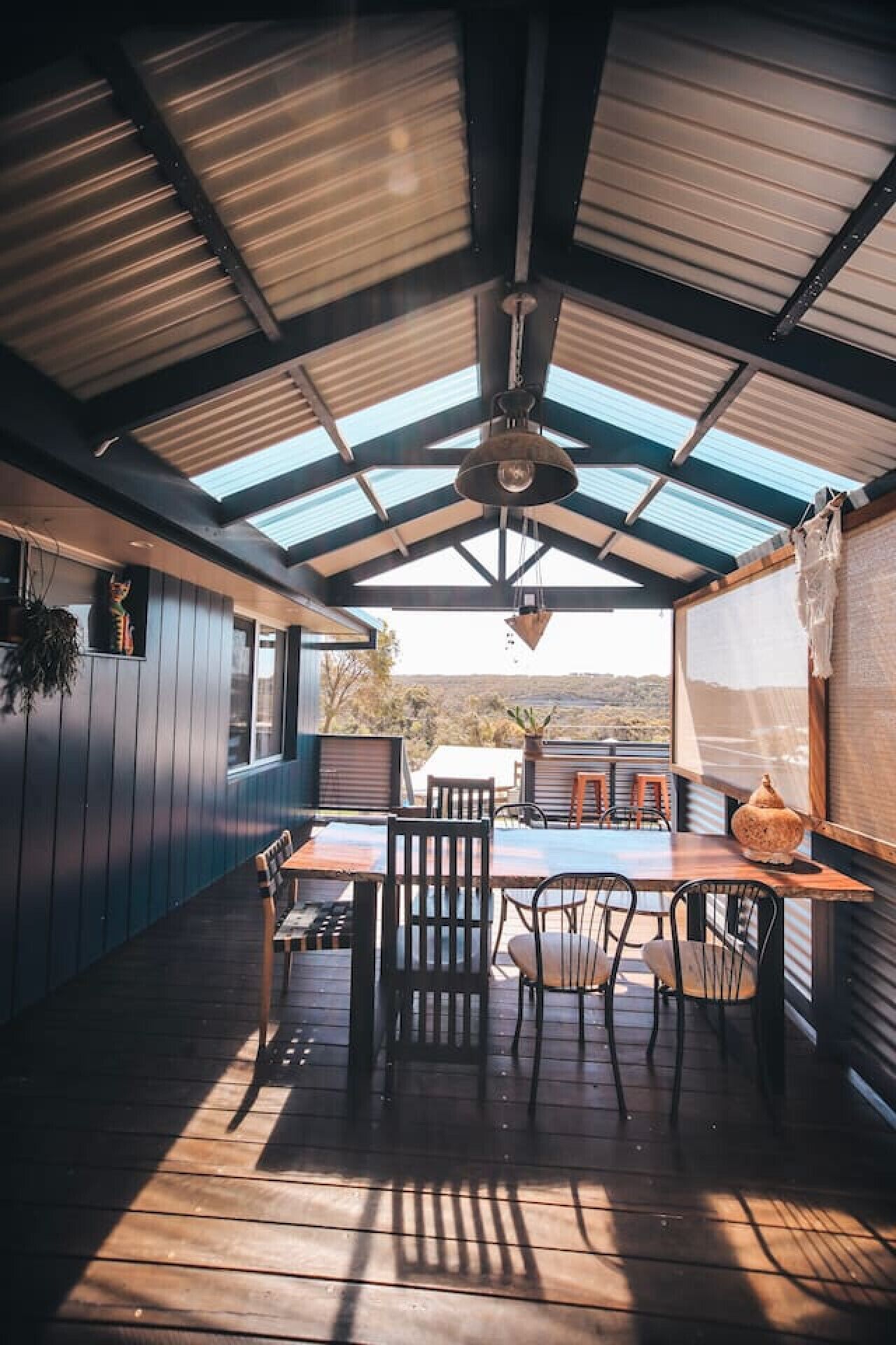 An outdoor patio with a wooden table and chairs, a hanging light fixture, and a view of the landscape.