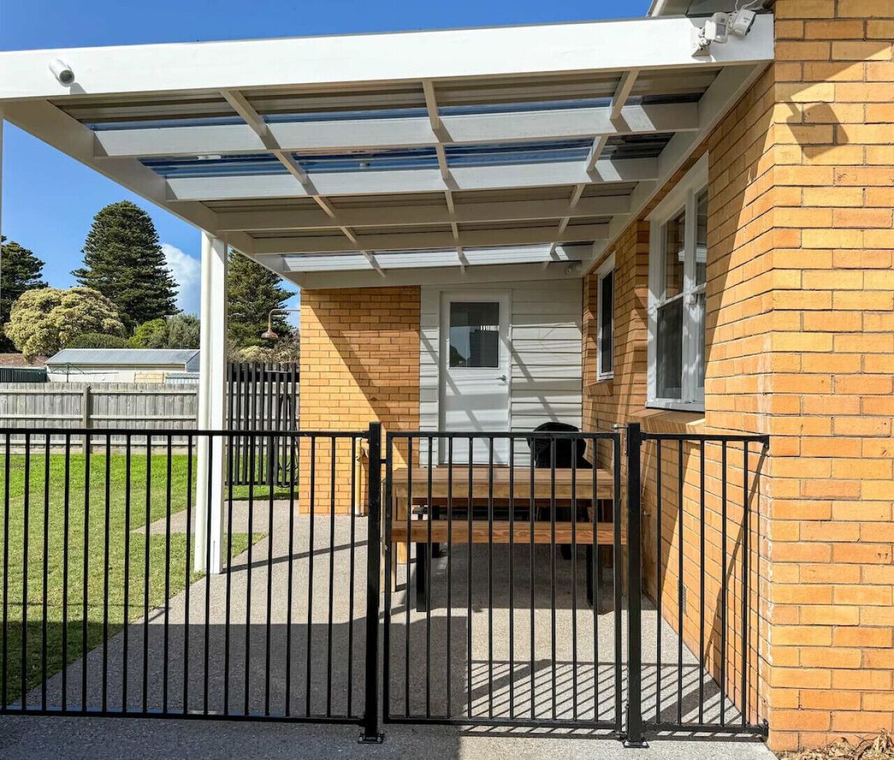A covered outdoor area with a metal gate, a wooden bench, and a brick building with a white door.