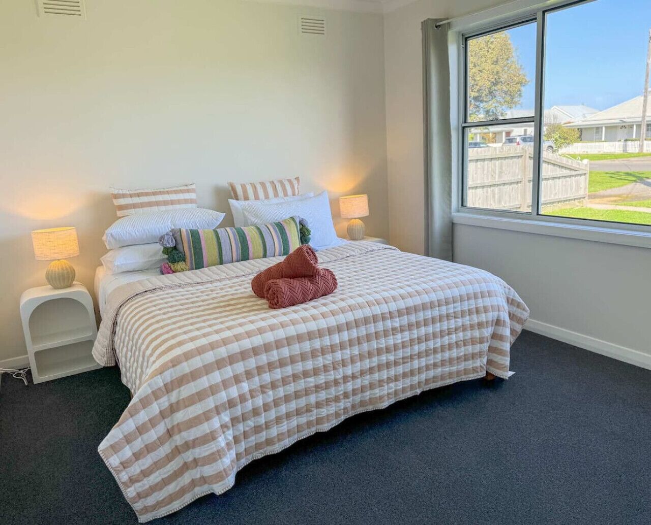 A bedroom with a bed, pillows, a nightstand, a lamp, and a window with a view of a residential area.