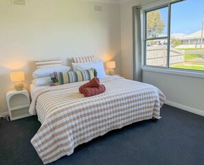 A bedroom with a bed, pillows, a nightstand, a lamp, and a window with a view of a residential area. - Campbell Cottage Port Fairy (Port Fairy)
