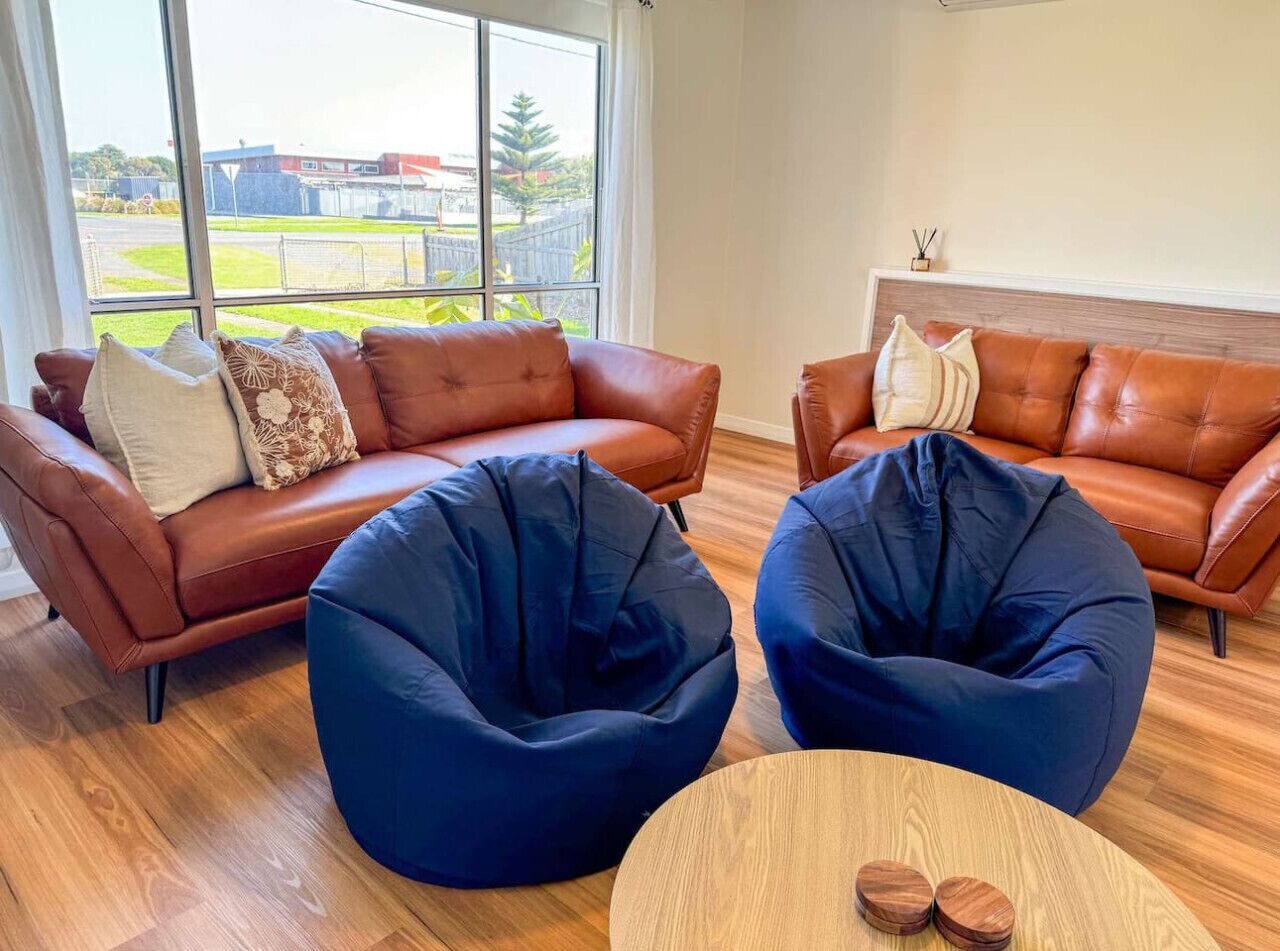 A modern living room with leather sofas, two bean bag chairs, a round wooden table, and a view of a green outdoor area through large windows.