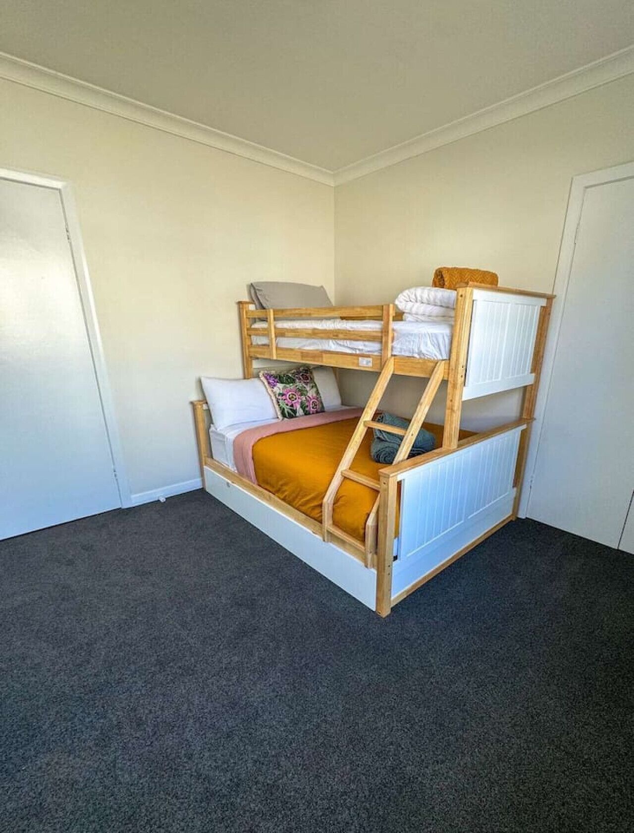Una cama individual con estructura de madera, un colchón y almohadas en una habitación con suelo alfombrado y paredes blancas.