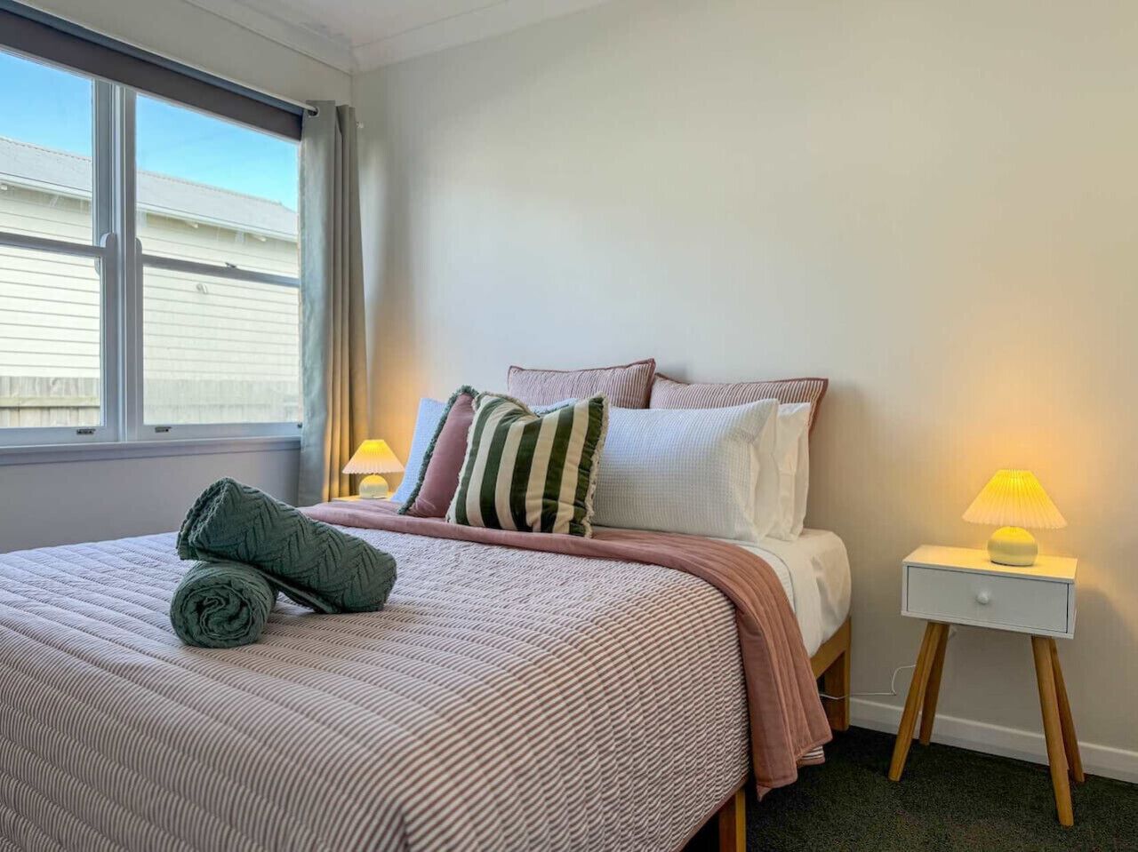 A neatly made bed with a striped bedspread, pillows, and a rolled towel. There is a bedside table with a lamp and a small drawer.