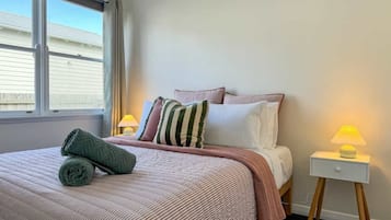 A neatly made bed with a striped bedspread, pillows, and a rolled towel. There is a bedside table with a lamp and a small drawer.