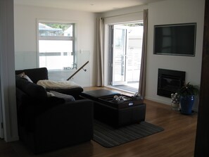A modern living room with a sofa, ottoman, and a flat-screen TV mounted on the wall. - Waihi Beach NORTH end and NO roads to cross (Waihi Beach)