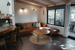 A modern living room with a brown leather sofa, a round wooden coffee table, and a kitchen area with a sink and bar stools.