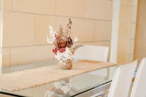 A glass table with a woven placemat, a vase with dried flowers, and white chairs.
