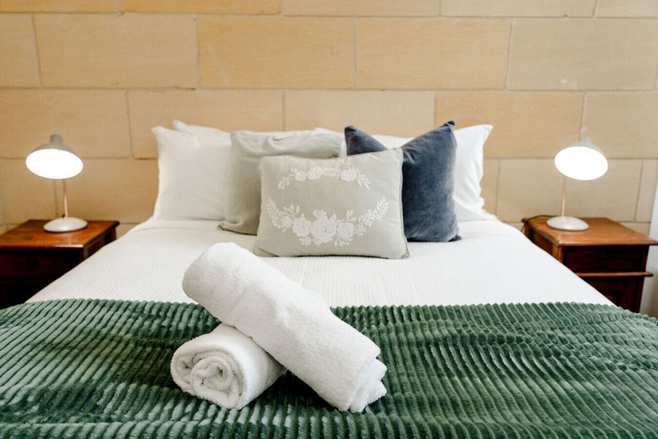 Una cama bien hecha con una colcha verde con textura, almohadas blancas y una toalla blanca enrollada sobre la cama.