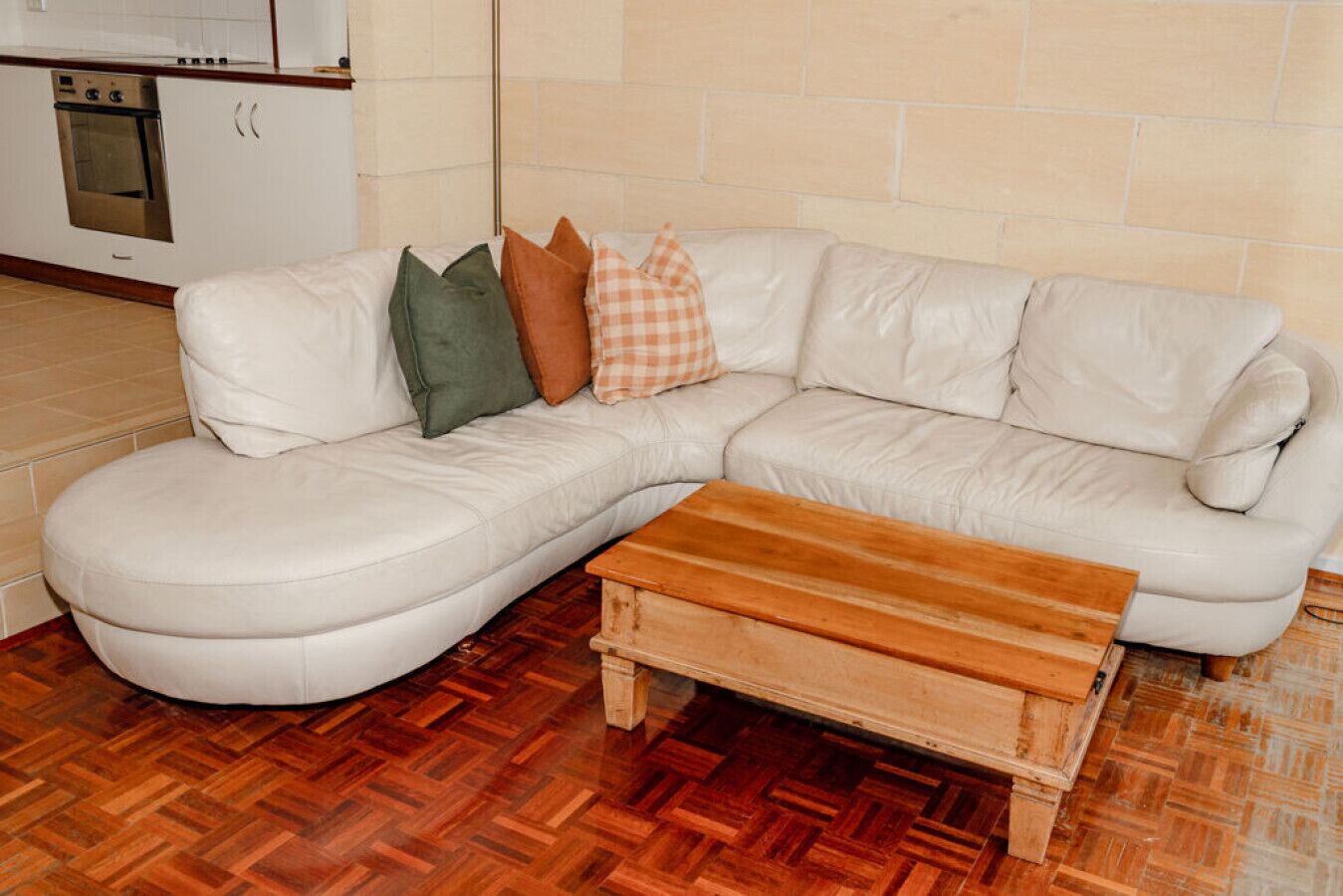 Un canapé d’angle en cuir blanc, accompagné d’une table basse en bois, dans une pièce dont le sol est recouvert de parquet à motifs en chevrons.