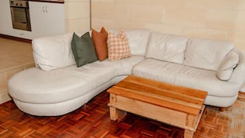 Un canapé d’angle en cuir blanc, accompagné d’une table basse en bois, dans une pièce dont le sol est recouvert de parquet à motifs en chevrons.