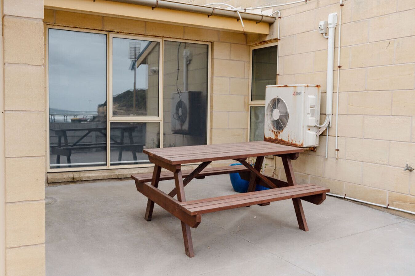 Une table de pique-nique en bois, dotée d’un banc intégré, est placée à l’extérieur d’un bâtiment aux grandes fenêtres, sur le mur duquel est fixée une unité de climatisation.