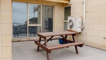 Une table de pique-nique en bois, dotée d’un banc intégré, est placée à l’extérieur d’un bâtiment aux grandes fenêtres, sur le mur duquel est fixée une unité de climatisation.