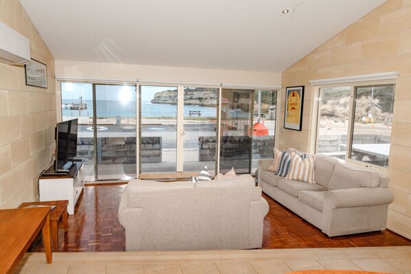 A living room with a sofa, a television, and a view of the sea through large glass doors.
