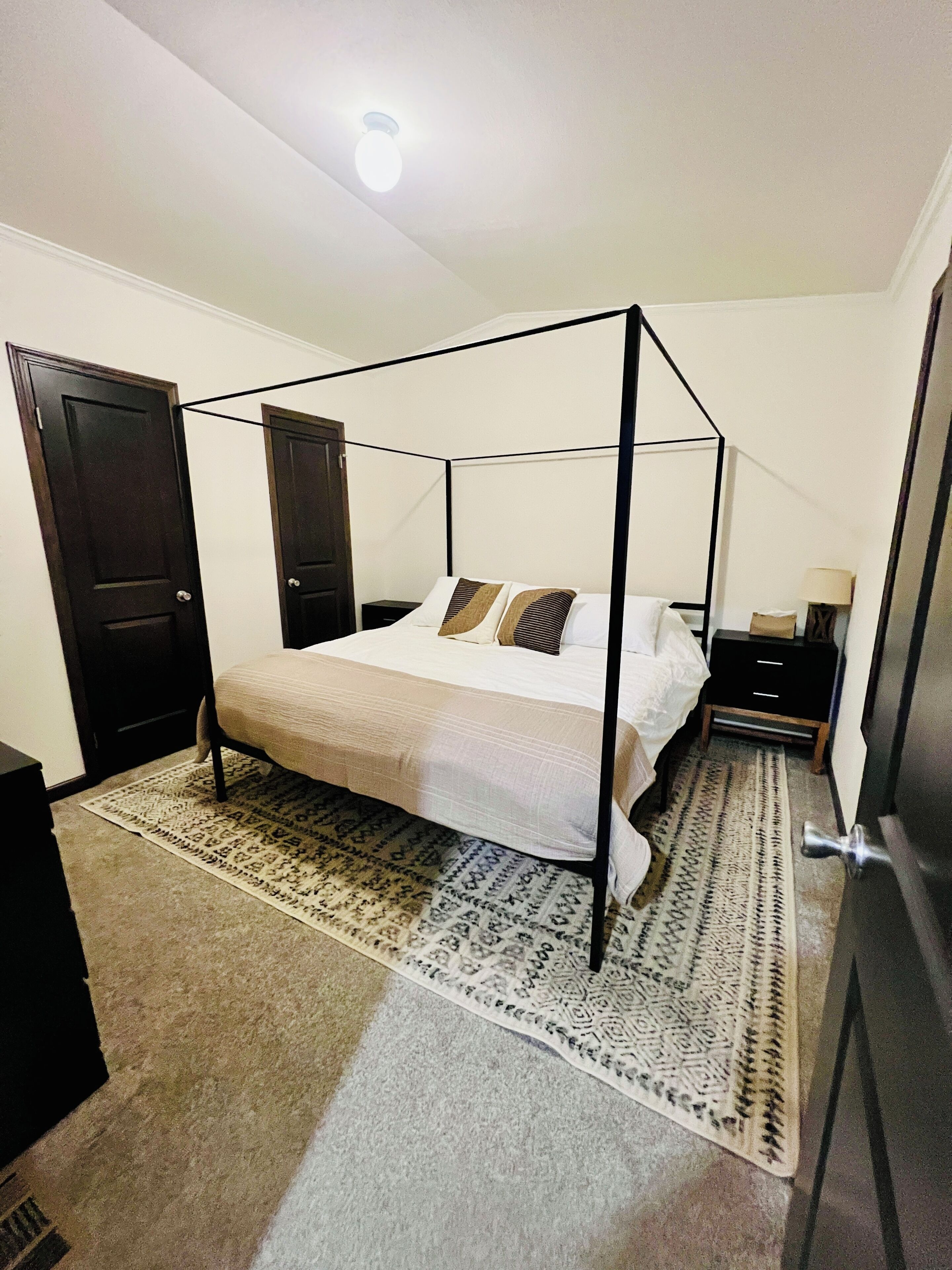 Ein Bett mit Baldachin, mit weißer Bettdecke und zwei Kissen, ein Nachttisch mit Lampe sowie ein gemustertes Teppich auf einem Teppichboden.
