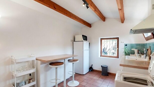 A compact kitchen with a wooden ceiling, a white refrigerator, a window with a view, and a small stool. - Charmante maisonnette (La Rochelle)